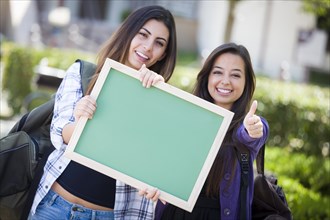 Portrait of two attractive mixed race female students holding blank chalkboard with thumbs up and carrying backpacks on school campus