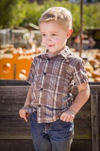 Adorable little boy at pumpkin patch with hands in his pockets leaning on antique wood wagon in rustic ranch setting