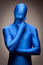 Thinking man with hand on chin wearing full blue nylon bodysuit on a grey background