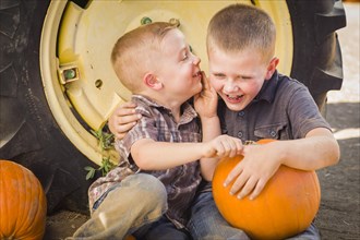 Two boys sitting against a tractor tire holding pumpkins and whispering secrets in rustic setting