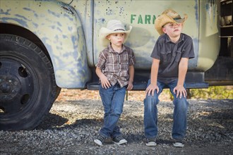 Two young boys wearing cowboy hats leaning against an antique truck in a rustic country setting