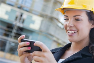 Young professional female contractor wearing hard hat at contruction site texting with cell phone