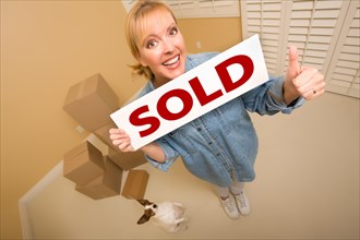 Excited woman with thumbs up and doggy holding sold real estate sign near moving boxes in empty room taken with extreme wide angle lens