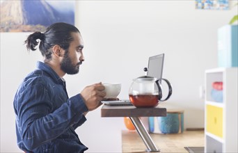 Young man with ponytail and checkered shirt drinking tea at home and working