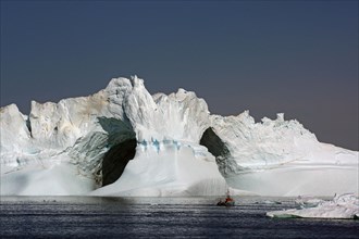 View of small fishing boat in front of huge iceberg with 2 holes