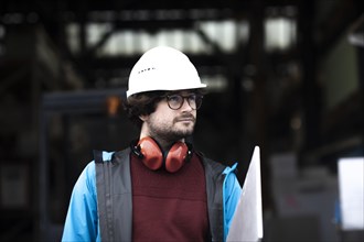 Young engineer with helmet and hearing protection checks with tile in front of a warehouse