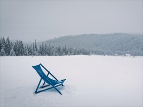 Sunbed type chair on the top of a mountain in a snowy winter day. Fir forest covered with snow on the horizon. Blizzard scene