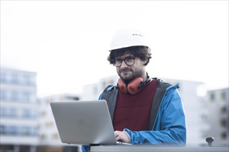 Young engineer with helmet and hearing protection checks outside work with laptop