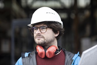 Young engineer with helmet and hearing protection checks with tile in front of a warehouse