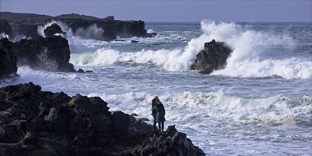 Two people standing embraced on the black lava coast looking out over the stormy surf