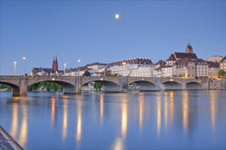 View from the banks of the Rhine along the river promenade to the old town of Basel illuminated at night with the Basel Cathedral