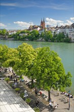 View from the river promenade of Basel Cathedral and the old town of Basel with the turquoise Rhine River and the lively river promenade in the foreground