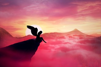 Silhouette of a lonely fallen angel with long wings standing on a cliff against a paradise sunset