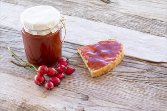 Homemade rosehip jam in a jar and slice of bread with spread on wooden table with ripe fruit