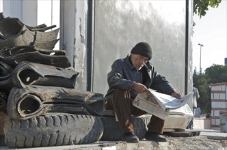Man with cap sits next to cut car tyres and reads newspaper