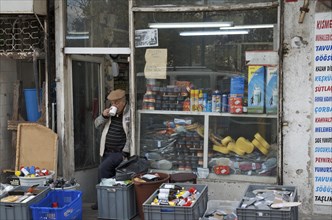 Old man drinking tea in front of his shop with consumer goods