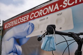 A bicycle with a surgical mask on the handlebars stands on a large poster in front of a Corona rapid test station