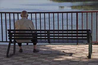 Old man sitting alone on park bench by river in front of bridge