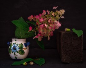 Still Life with Hydrangea Blossom in Colourful Ceramic Vase Next to Wooden Block and Acorns