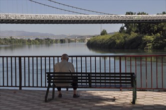 Old man sitting alone on park bench by river in front of bridge
