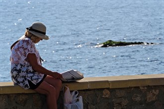 Woman with shopping bags sitting on waterfront reading newspaper