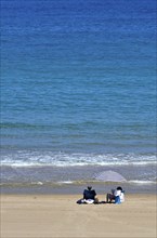 Man and woman under parasol on beach at water's edge from sea