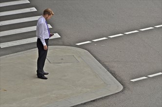 Man from office stands at zebra crossing and looks at his mobile phone