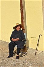 Old woman with straw hat and walking stick sitting in front of yellow house wall