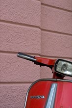 Handlebars and front of a red scooter in front of a pink house wall