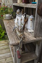 Old workbench decorated with antique tea kettles and manual meat grinder stuffed with old wrenches on wooden deck behind old house