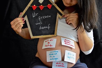 Expectant with wish slips pasted on her stomach holds up black board with saying Wir freuen uns drauf