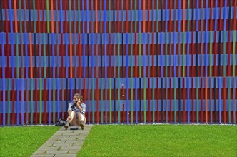 Young woman on the phone sitting on the floor in front of the colourful striped facade of the Museum Brandhorst