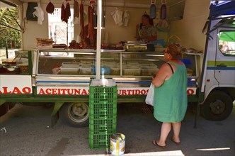 Fat woman in airy dress stands in front of sales cart with olives