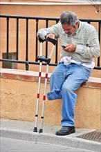 One-legged man with crutch sits on wall and looks into his mobile phone