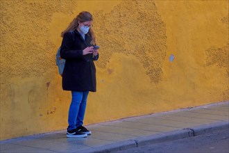 Young woman with mobile phone and mouth guard in front of yellow wall in La Chanca district