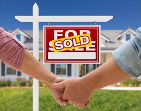 A couple hold his hands in front of a beautiful new house with sign for sale