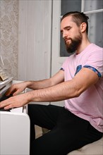 A young man with a well-groomed long beard and a pink shirt sits at the piano and plays music