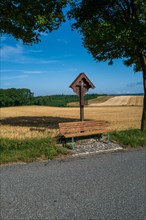A wayside cross with a park bench at the edge of a wheat field in summer