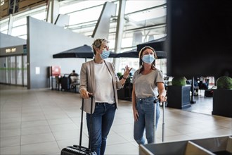 Two travelling women wearing protective masks discussing by flight information board at the Faro airport