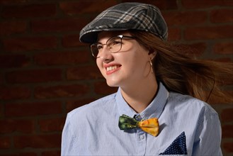 Pretty very young woman with long brunette hair wears a blue shirt with pocket square and bowtie and a checked cap. The smile with open mouth