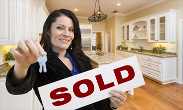 Pretty hispanic woman in kitchen holding house keys and sold sign