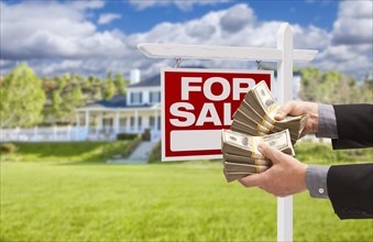 Man handing over thousands of dollars in front of house and for sale real estate sign