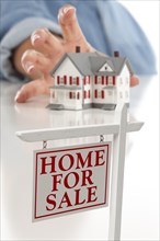 Real estate sign in front of womans hand reaching for model house on a white surface