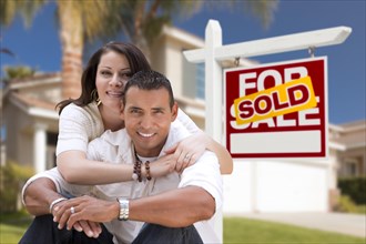 Young happy hispanic young couple in front of their new home and sold for sale real estate sign