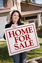 Hispanic woman holding home for sale real estate sign in front of house