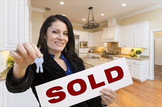 Pretty hispanic woman in kitchen holding house keys and sold sign