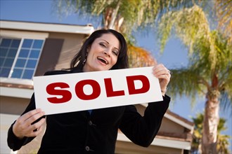 Happy attractive hispanic woman holding sold sign in front of house