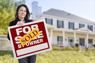 Smiling hispanic female holding sold for sale by owner sign in front of beautiful house