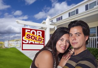 Young happy hispanic couple in front of their new home and sold for sale real estate sign