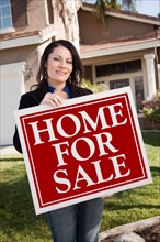 Happy attractive hispanic woman holding home for sale real estate sign in front of house